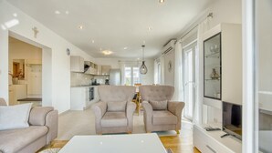 Ein modernes Wohnzimmer mit zwei Sofas, einem Couchtisch und einem Fernseher. Im Hintergrund befindet sich eine Küche mit weißen Schränken und einem Essbereich.