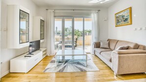 Ein modernes Wohnzimmer mit Fernseher, einer Couch, einem Glastisch und Blick ins Freie durch große Glasfenster.