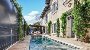 Un espace piscine doté d’une terrasse en bois, d’une pergola et d’un bâtiment à la façade en pierre et aux volets bleus.