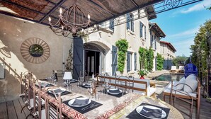 Un espace de restauration en plein air avec une longue table dressée pour un repas, un lustre et une vue sur une piscine et un bâtiment en arrière-plan.