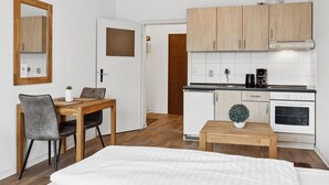 A compact hotel room with a bed, a small dining table with two chairs, a kitchenette with a stove, a coffee maker, and a potted plant.