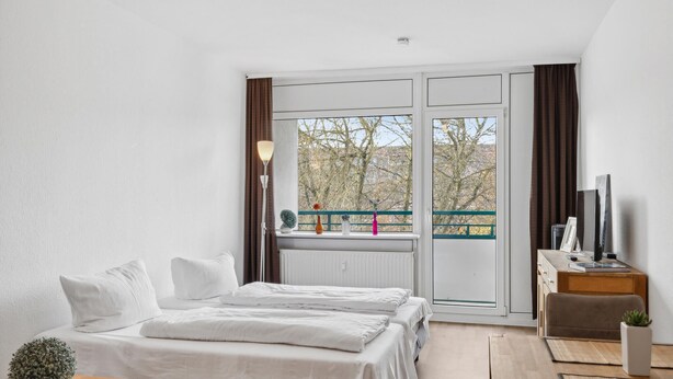 A hotel room with a bed, a desk, a chair, a small table, and a balcony with a view of trees.