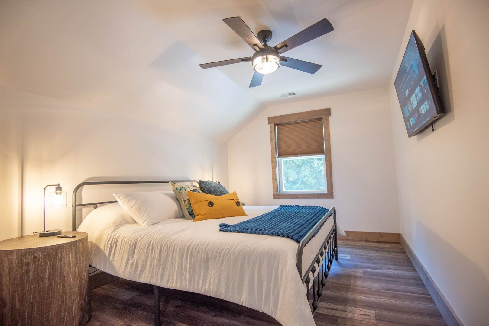 A bedroom with a bed, a ceiling fan, a TV mounted on the wall, and a window with blinds.