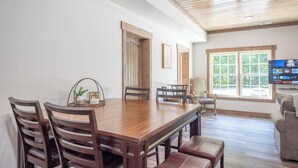 A dining area with a wooden table and chairs, a television mounted on the wall, and a window with a view of greenery.