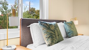 A neatly made bed with white and blue pillows, a bedside lamp, and a view of trees and a building outside the window.