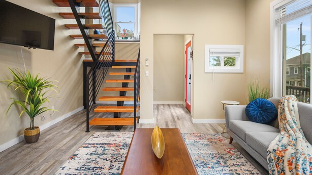 A modern living room with a wooden staircase, a flat-screen TV mounted on the wall, a sofa with a blue pillow, and a patterned rug.