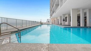 Una piscina con barandilla de metal, cielo azul despejado y un edificio con varios balcones.