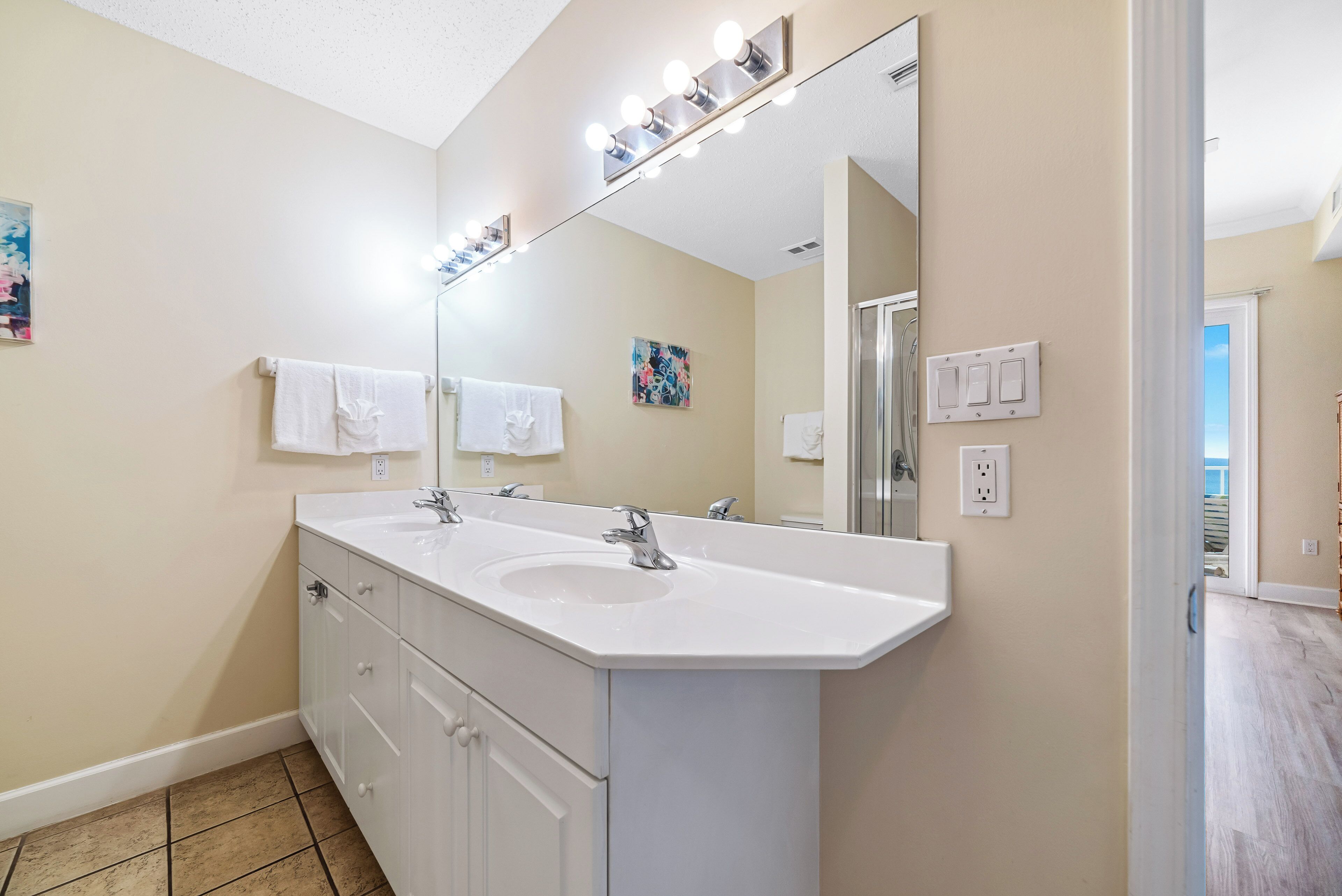 A bathroom with a double sink vanity, a large mirror with lights, and a painting on the wall.