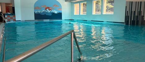 An indoor swimming pool with a mural of mountains, large windows, and a metal handrail.