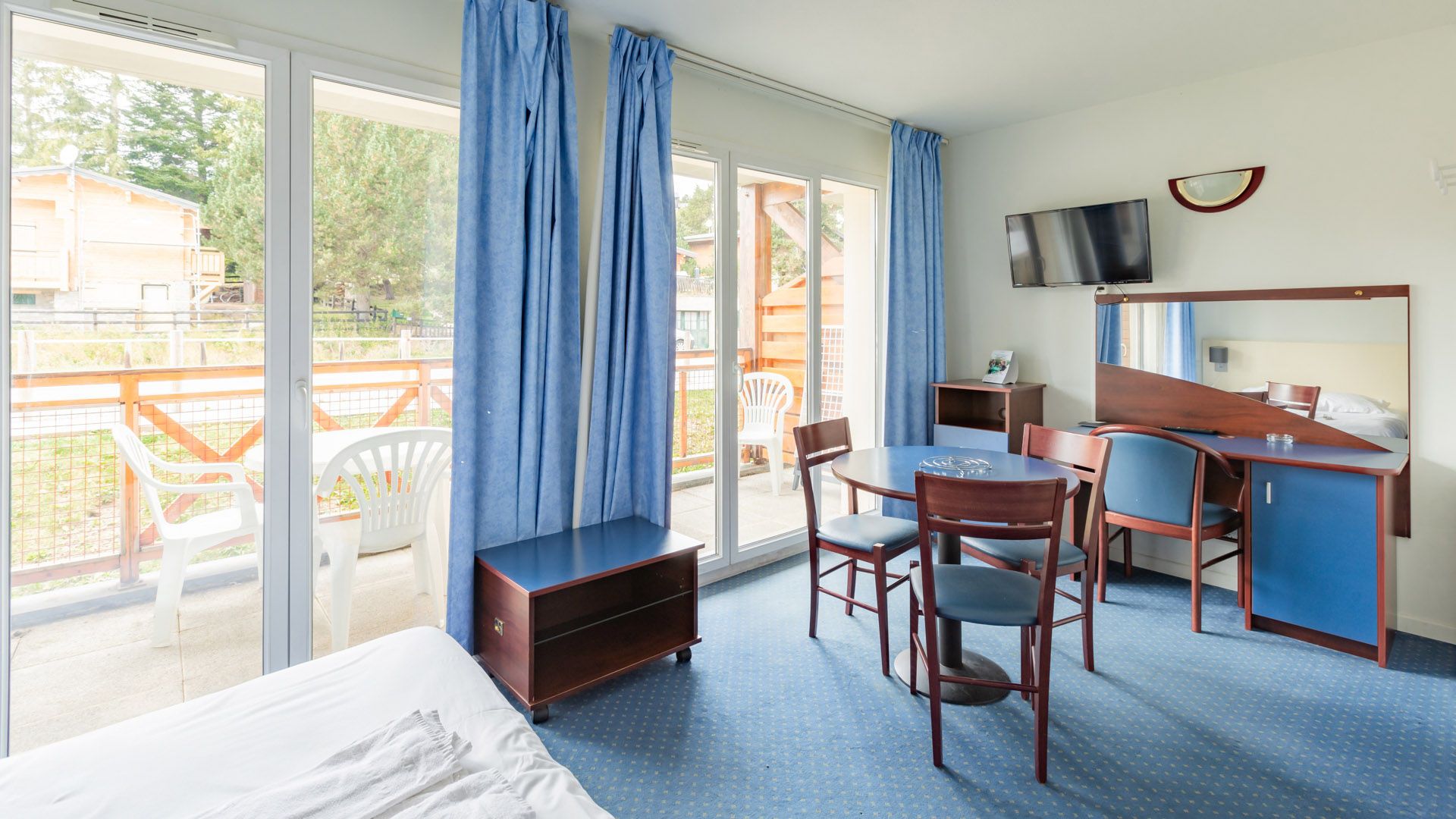 A hotel room with a bed, a desk, a chair, a TV, and a balcony with a view of the outside.