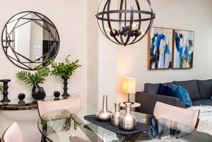 A modern living room with a glass dining table, a geometric mirror, abstract art, and a dark-colored sofa.