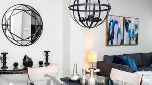 A modern living room with a glass dining table, a geometric wall mirror, abstract art, and a spherical ceiling light fixture.
