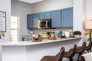 A modern kitchen with blue cabinets, a stainless steel microwave, a coffee maker, and a stove. There are wooden bowls and a plant on the counter.