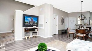A modern living room with a flat-screen TV, a dining area, and a staircase.