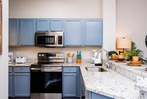 A compact kitchen with blue cabinets, a stainless steel microwave, a built-in oven, a sink, and a countertop with a speckled pattern.
