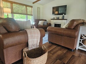 A living room with two brown leather sofas, a wicker basket, a fireplace, and a mounted TV. - The Monarch - Private Setting, Heated Pool, 6 Bedroom, New Build. 6 min to CDP (Fly Creek)
