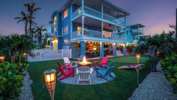 A two-story building with a balcony, outdoor seating area with chairs and a fire pit, surrounded by palm trees and a well-maintained lawn. - Anna Maria Island of Palms - HotTub,Pool,Poker,Games and more (ANNA MARIA)
