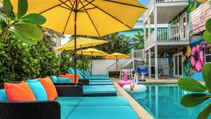 A poolside area with lounge chairs and umbrellas, a pool, and a building with a colorful mural. - Anna Maria Island of Palms - HotTub,Pool,Poker,Games and more (ANNA MARIA)