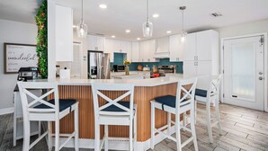 A modern kitchen with white cabinets, a central island with bar stools, pendant lighting, and a decorative plant wall. - Anna Maria Island of Palms - HotTub,Pool,Poker,Games and more (ANNA MARIA)
