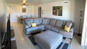 A living room with a grey sectional sofa, two yellow pillows, a grey ottoman, a ceiling fan, a framed picture, a dining table with chairs, and a floor lamp.