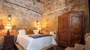 Een slaapkamer met een bakstenen muur, een houten garderobe, een bed met wit beddengoed en twee bedlampjes.