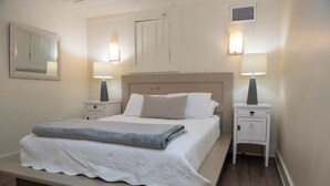 Un lit bien fait, recouvert d’une couette blanche, deux tables de chevet avec des lampes, un miroir au mur et une porte avec un interrupteur.