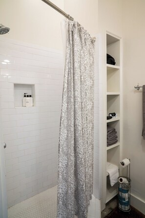 Eine Dusche mit gemustertem Vorhang, eine weiße Fliesenwand, ein Ablagefach mit Pflegeprodukten und ein Metallkorb mit Handtüchern.