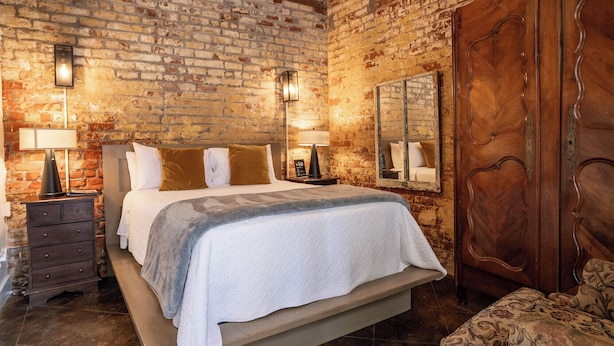 A room with a brick wall, a bed with a white and gray blanket, two bedside lamps, a wooden wardrobe, and a patterned chair.