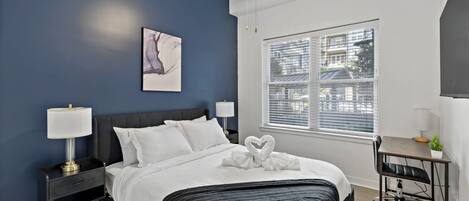 A modern bedroom with a dark blue accent wall, a large bed with white linens, a heart-shaped towel on the bed, a desk with a chair, and a window with blinds.