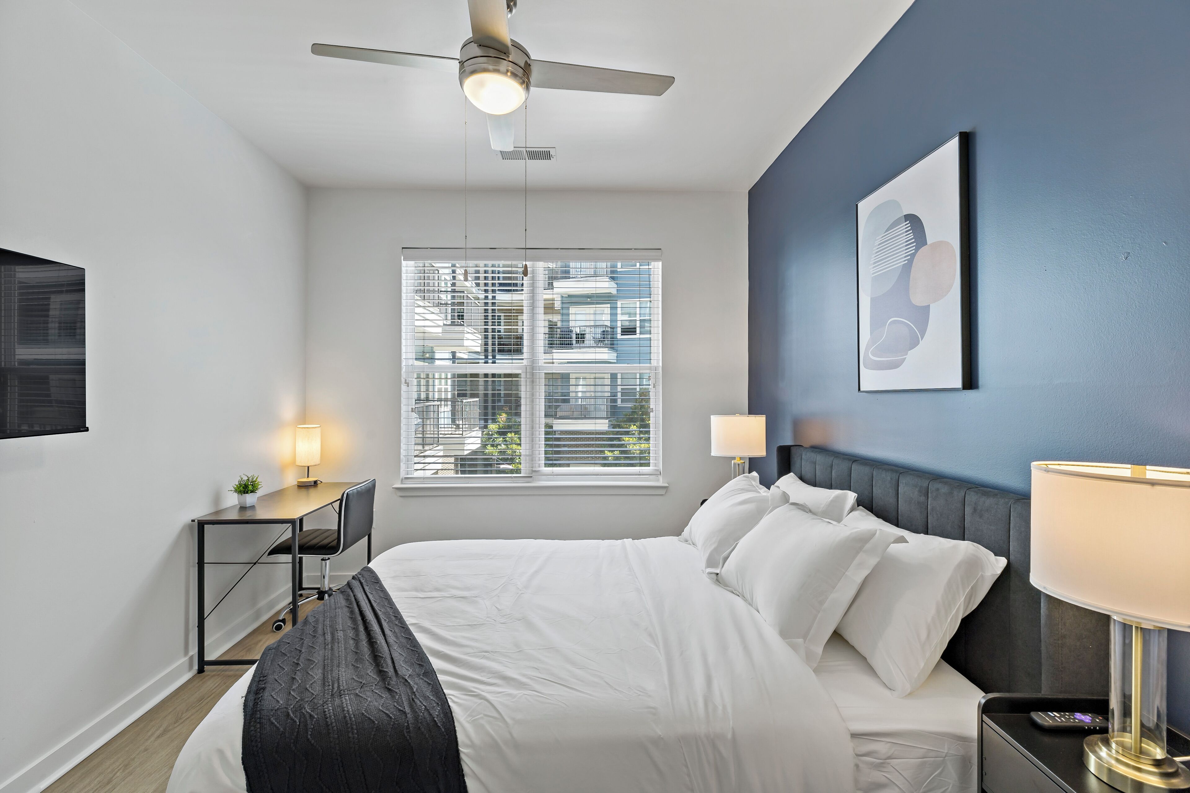 A modern hotel room with a bed, bedside lamps, a desk with a chair, a ceiling fan, and a window with a view of buildings.