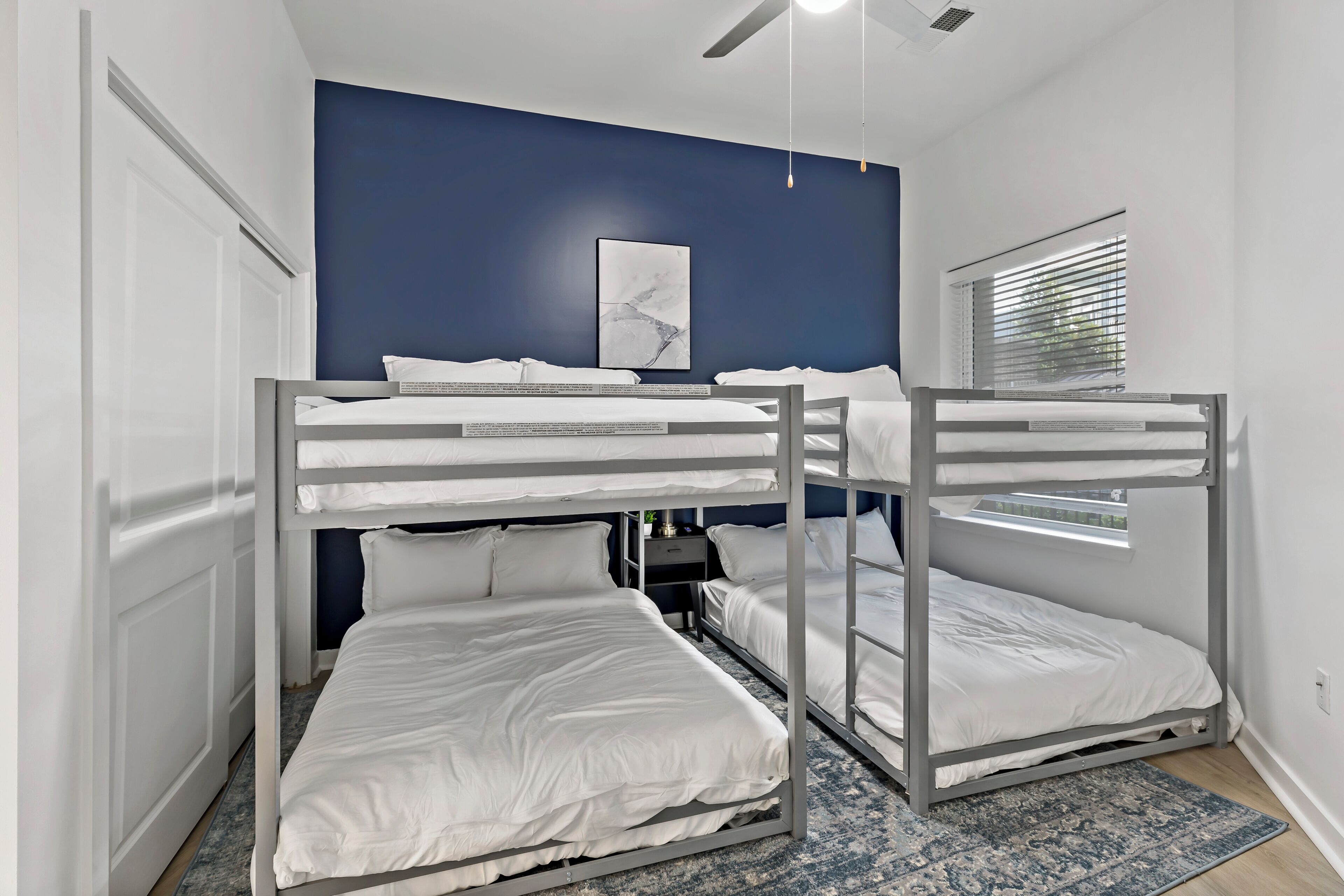 A bunk bed room with a blue accent wall, a ceiling fan, and a window with blinds.