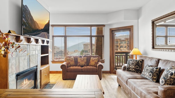 A modern living room with a fireplace, a large flat-screen TV, a brown leather sofa, and a wooden coffee table.
