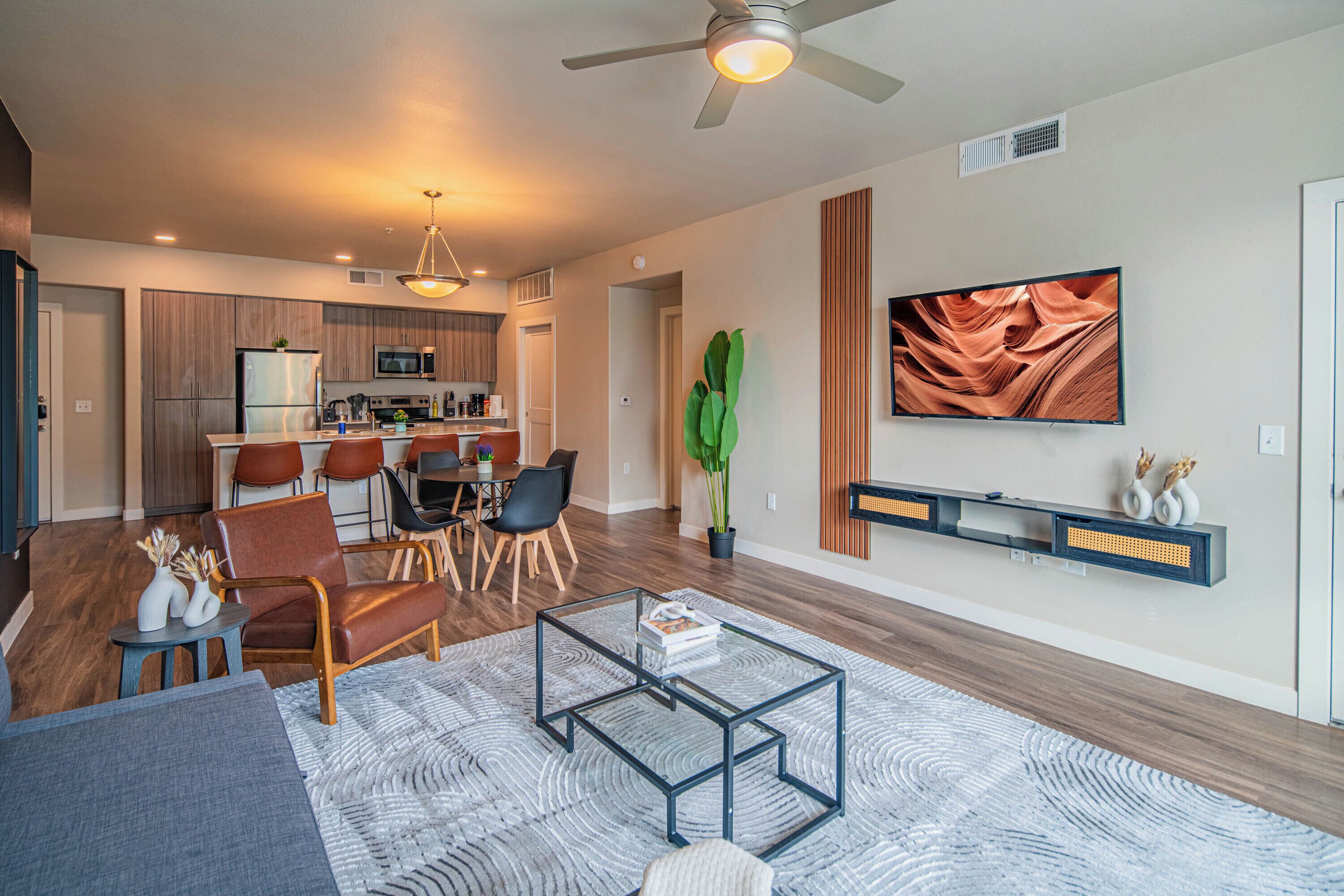 A modern living room with a glass coffee table, a dining area with chairs, a kitchen with wooden cabinets, and a mounted TV.