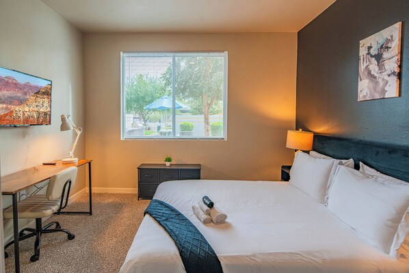 A hotel room with a large bed, a desk with a lamp, a small table with a plant, and a window with blinds. - ROVE LIVING/KB - V on Broadway (Tempe)