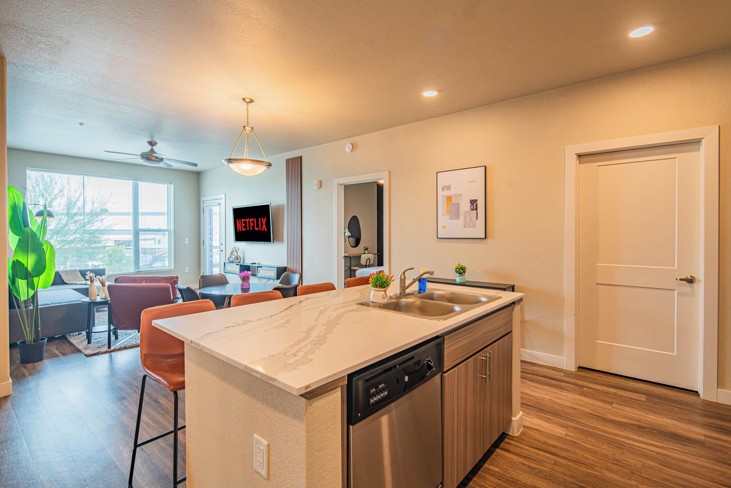 A modern kitchen with a dishwasher, a sink, and a dining area with a TV displaying 'NETFLIX'.