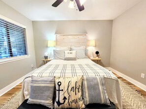 A bedroom with a bed, two bedside tables, a nightstand, a window with blinds, a ceiling fan, and a wall-mounted lamp.