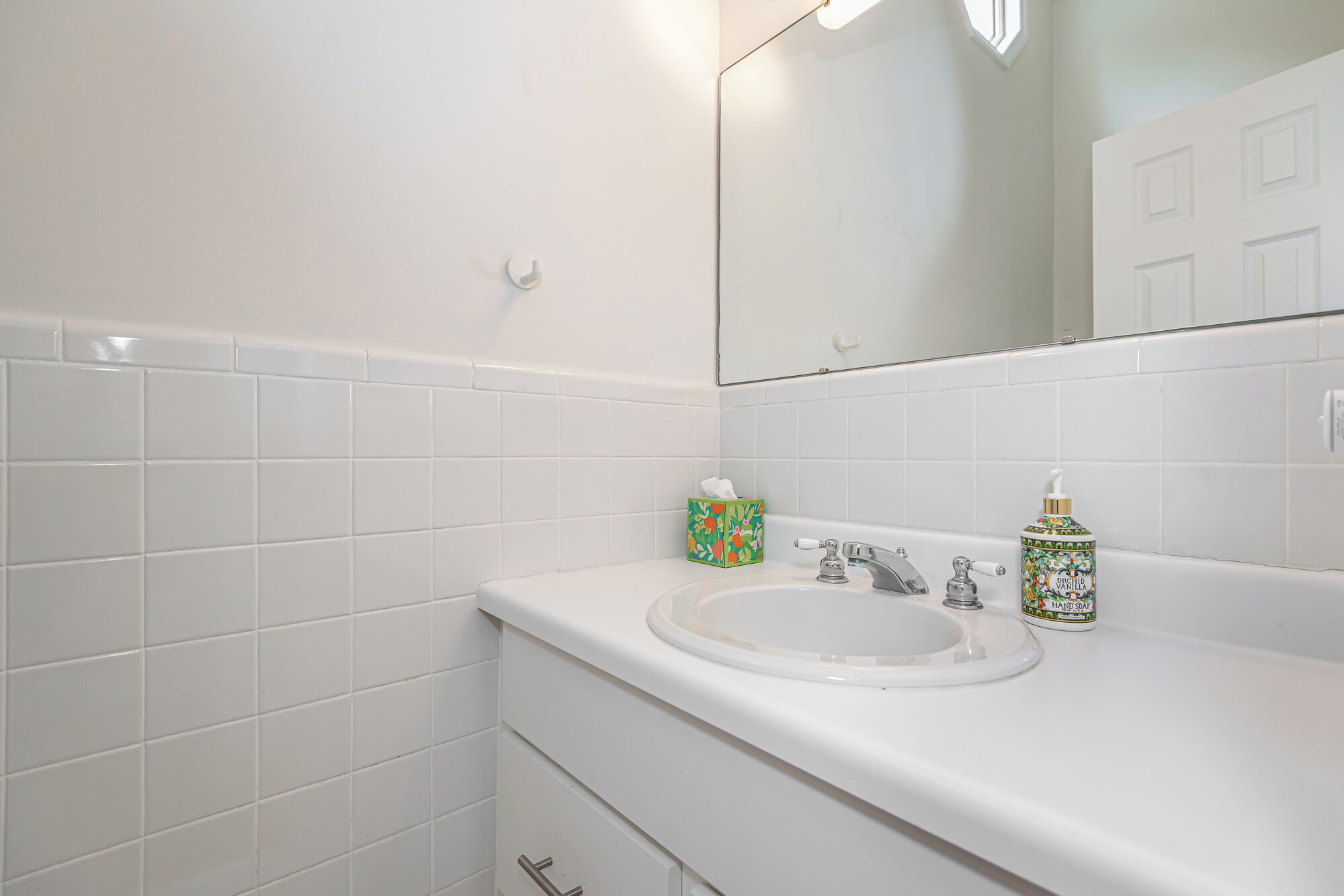 A bathroom with white tiled walls, a white sink, a mirror, and a hand soap dispenser.