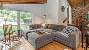 Una sala de estar acogedora con techo de madera, un sofá seccional gris, un sillón mecedor y una pared de ladrillo.