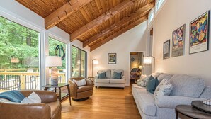 Amplia sala de estar con techo alto de madera, ventanas grandes y vistas al exterior. Hay dos sofás y una mesa con una lámpara.