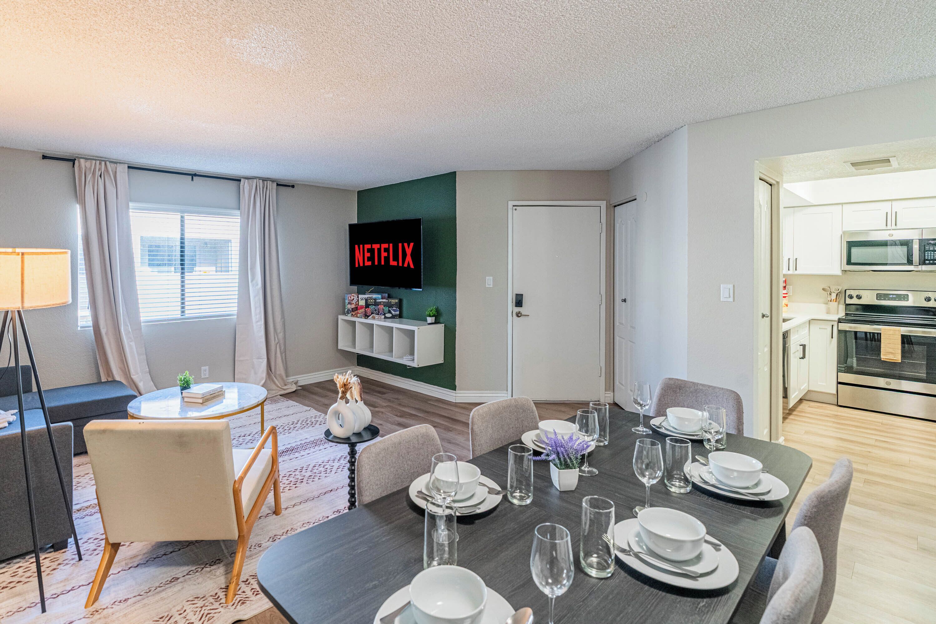A modern living room with a dining area, a flat-screen TV mounted on the wall, and a kitchen visible through an open door.