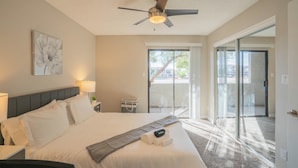 A hotel room with a bed, a ceiling fan, a window, and a sliding glass door leading to an outdoor area.