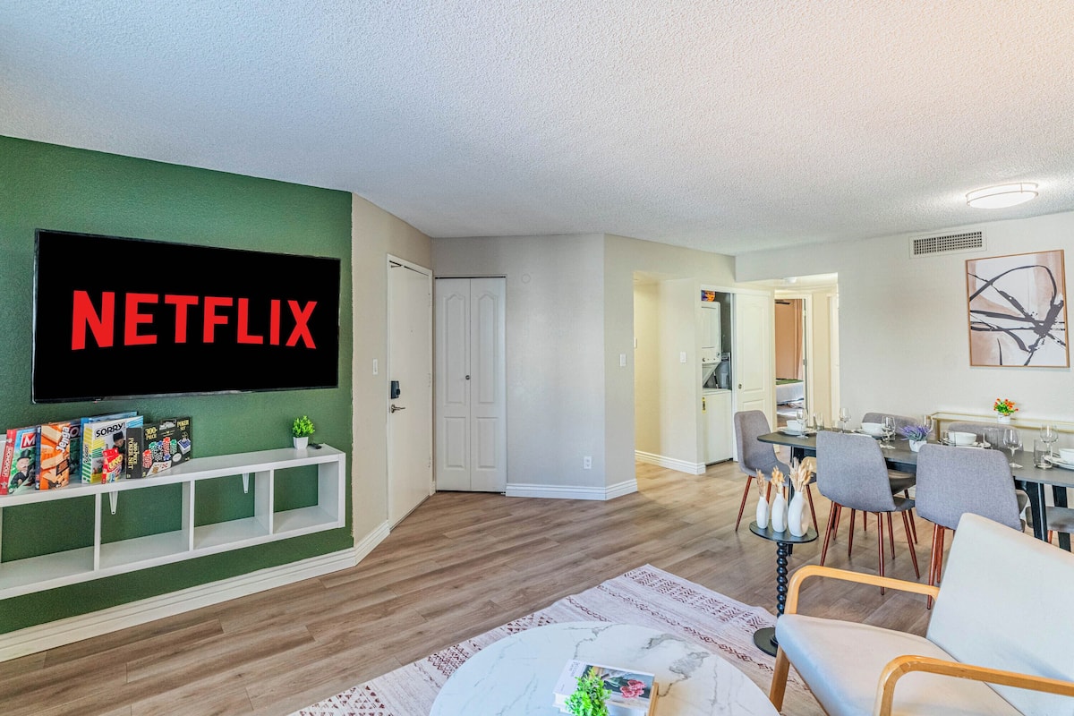 A modern living room with a flat-screen TV mounted on a green wall, a dining area with a table and chairs, and a hallway leading to a kitchen.