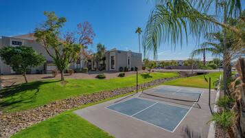 A residential area with a tennis court, a building with a 'Now Leasing' sign, and a clear blue sky.