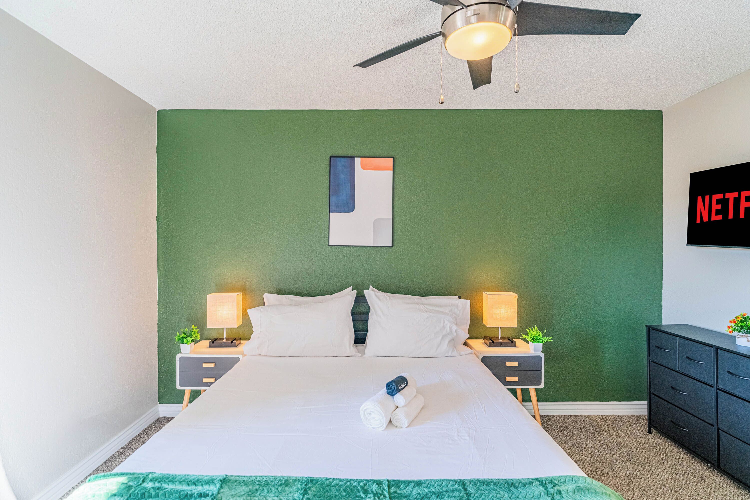 Una habitación de hotel con una pared verde de acento, una cama con ropa de cama blanca, dos mesitas de noche con lámparas, un ventilador de techo y un televisor que muestra el logo de Netflix.