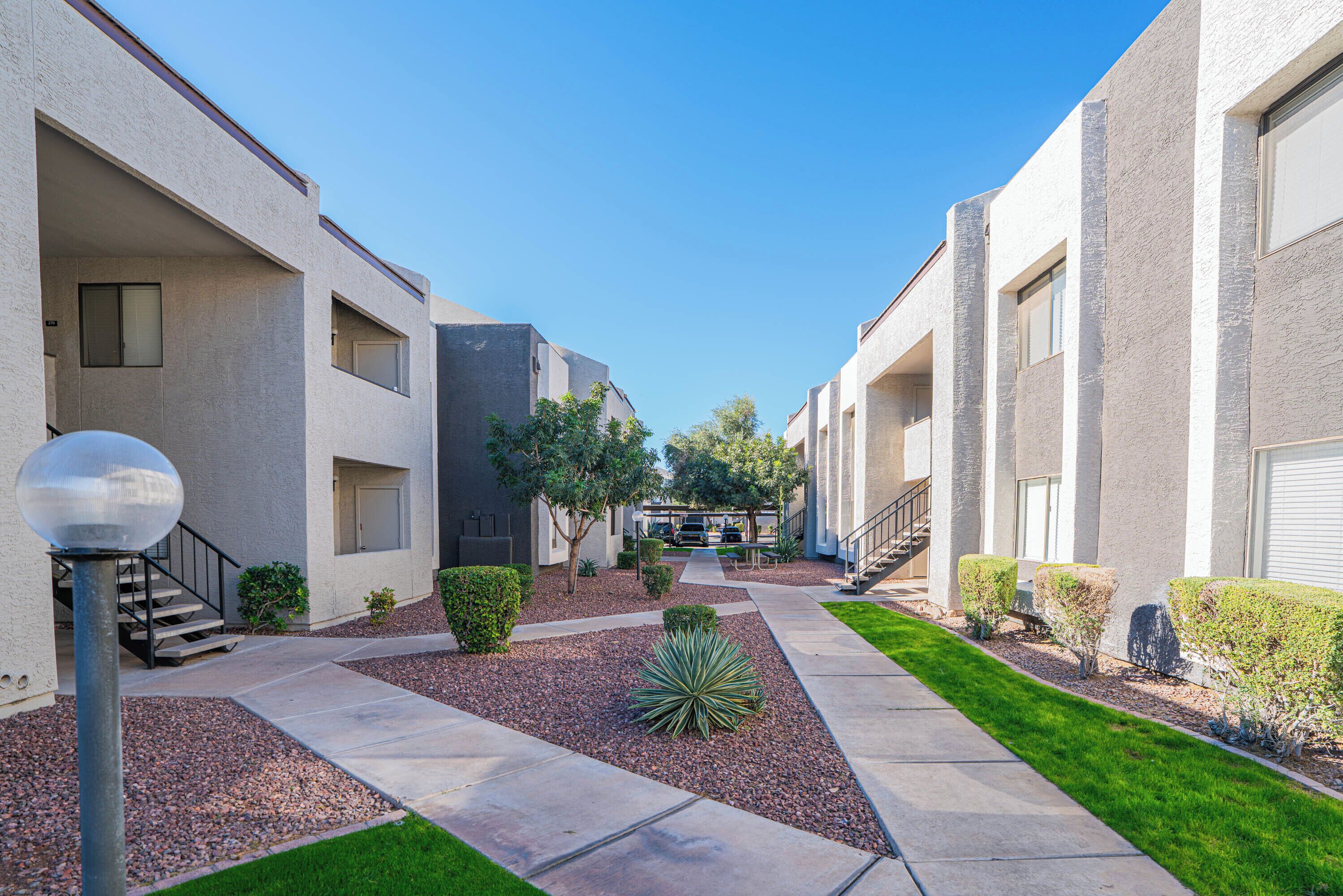 A modern apartment complex with a central courtyard, featuring a mix of single and double-story buildings, a well-maintained lawn, and a clear sky.