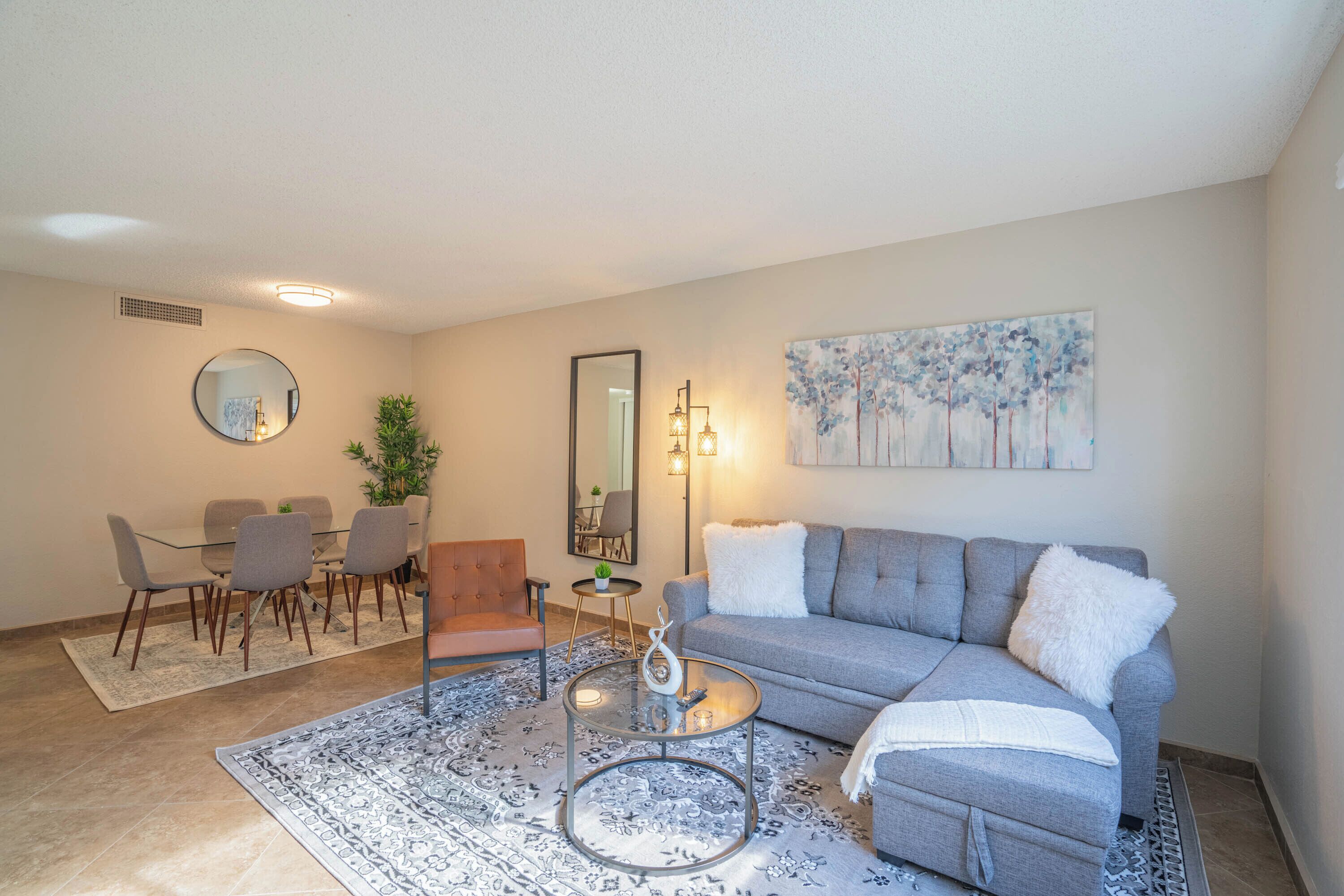 Uma sala de estar moderna com um sofá cinzento, mesa de centro redonda e um espaço de jantar com cadeiras e mesa.