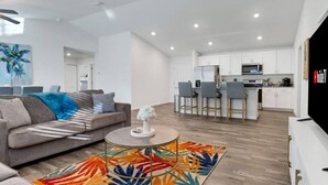 Ein modernes Wohnzimmer mit einer Couch, Couchtisch und einem farbenfrohen Teppich. Im Hintergrund ist eine Küche mit weißen Schränken und Barhocker erkennbar.