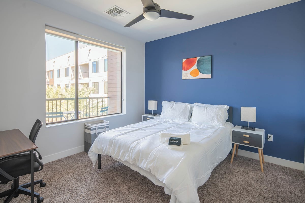 A bedroom with a large bed, a desk, and a chair. There is a window with a view of buildings outside.
