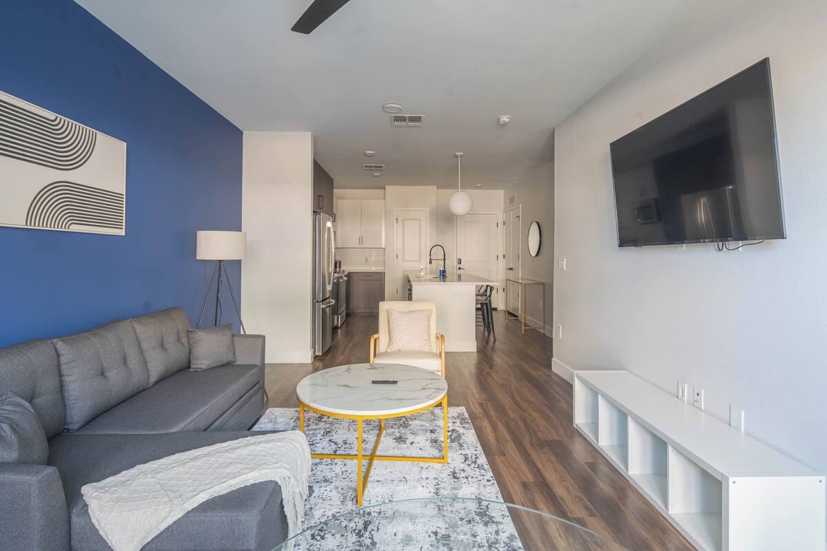 A modern living room with a grey sofa, a round coffee table, and a white side table. A kitchen with white cabinets and a dining area with a white chair are visible in the background.