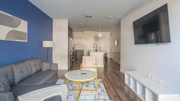 Una sala moderna con un sofá gris, una mesa de centro redonda y una mesita auxiliar blanca. Se ve una cocina con armarios blancos y un comedor con una silla blanca al fondo.
